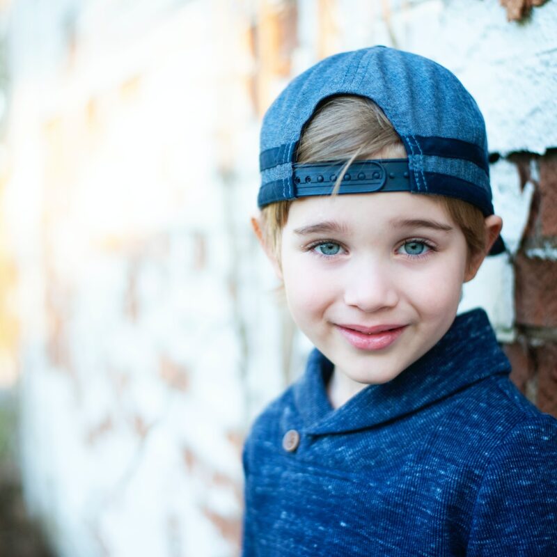 little boy with hat and blue sweater standing against brick wall