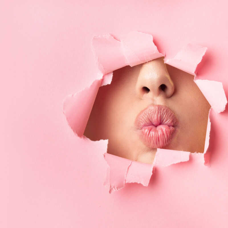 Unrecognizable Girl Pouting Lips Through Hole In Pink Paper. Makeup, Beauty Background. Panorama, Copy Space