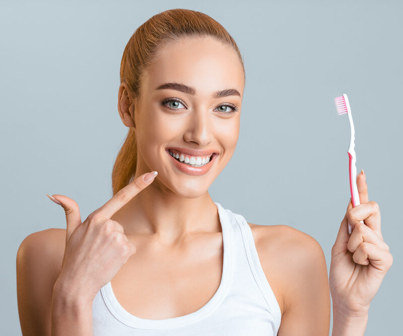 woman with slicked back blonde pony tail pointing to her teeth, holding a toothbrush in the other hand, wearing a white tanktop