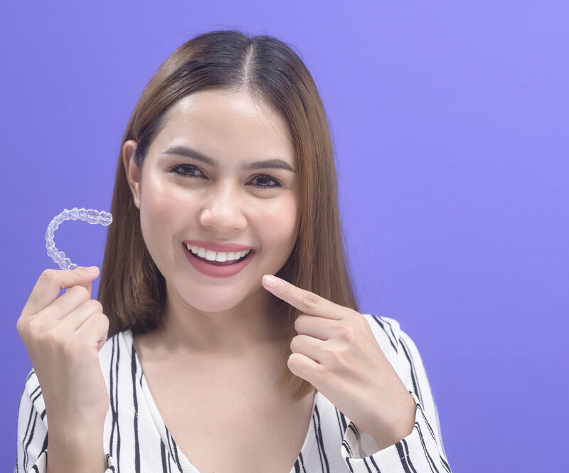 young woman holding aligners from orthodontist in one hand and pointing to her smile with the other on purple background