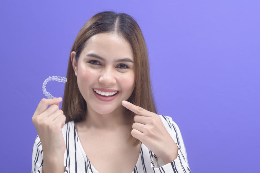 young woman holding aligners from orthodontist in one hand and pointing to her smile with the other on purple background